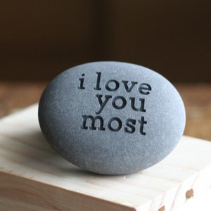 May include: A grey stone with the words "i love you most" engraved on it. The stone is sitting on a wooden surface.