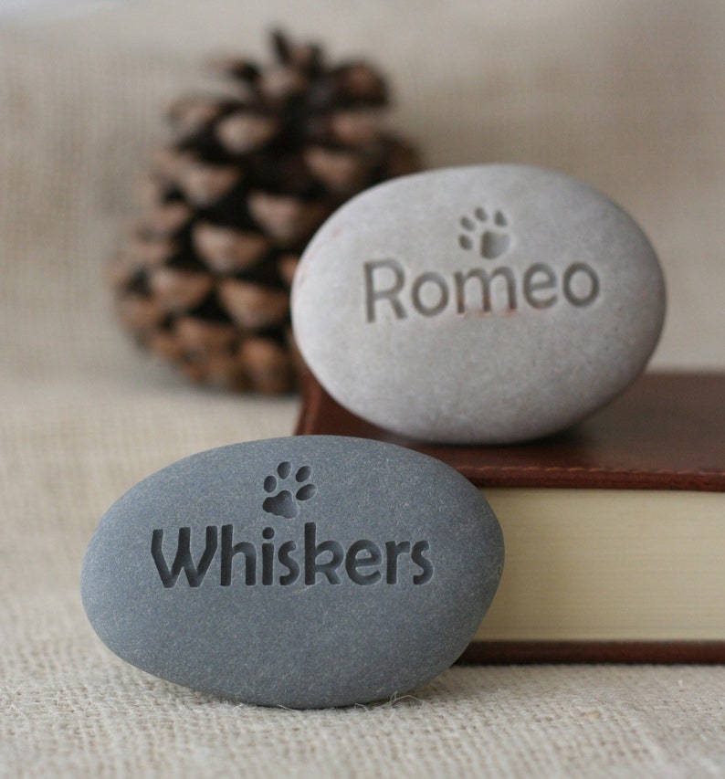 May include: Two gray stone pet memorial rocks with paw prints and the names "Romeo" and "Whiskers" engraved on them.