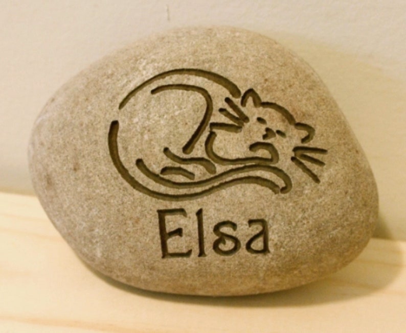 May include: A light brown stone with a carved image of a cat sleeping and the name "Elsa" engraved below.