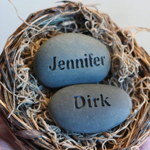 May include: Two grey stones with the names "Jennifer" and "Dirk" engraved on them, nestled in a brown bird's nest.