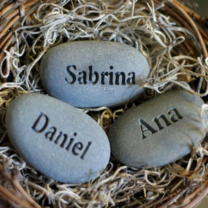 May include: Three grey stones with the names Sabrina, Daniel, and Ana engraved on them. The stones are nestled in a woven basket filled with dried moss.