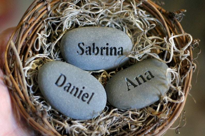 May include: Three grey stones with the names Sabrina, Daniel, and Ana engraved on them. The stones are nestled in a woven basket filled with dried moss.