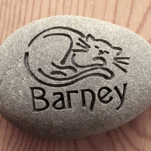 May include: A gray stone with a black engraved image of a cat sleeping and the name "Barney" underneath.