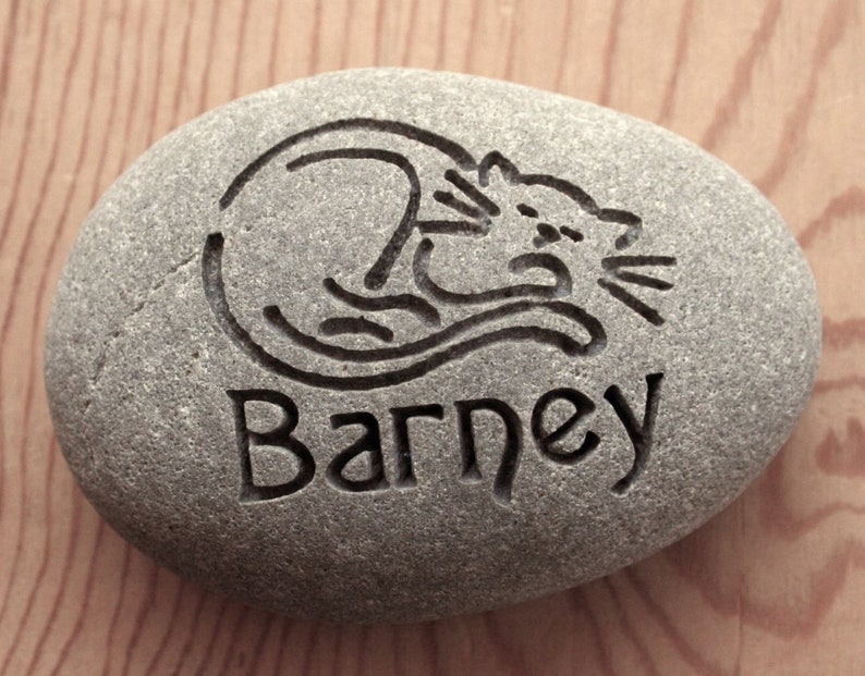 May include: A gray stone with a black engraved image of a cat sleeping and the name "Barney" underneath.