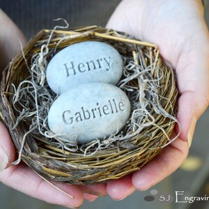 May include: Two grey stone eggs with the names "Henry" and "Gabrielle" engraved on them, nestled in a brown bird's nest.