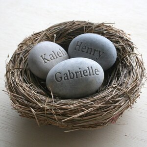May include: Three grey stones with the names "Caleb", "Henry", and "Gabrielle" engraved on them are nestled in a brown woven bird's nest.