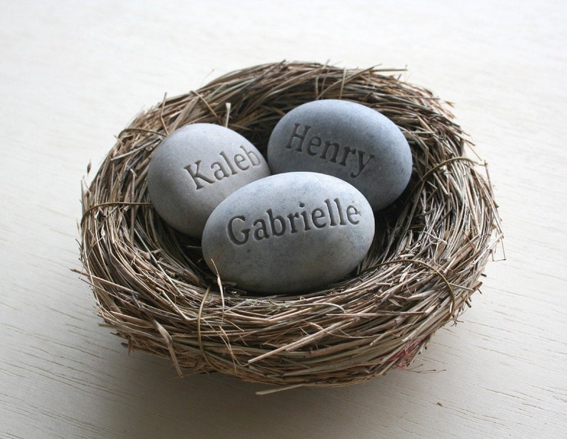 May include: Three grey stones with the names "Caleb", "Henry", and "Gabrielle" engraved on them are nestled in a brown woven bird's nest.