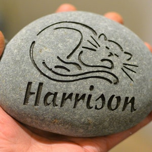 May include: A gray stone with a black etched design of a cat and the name "Harrison" engraved on it.