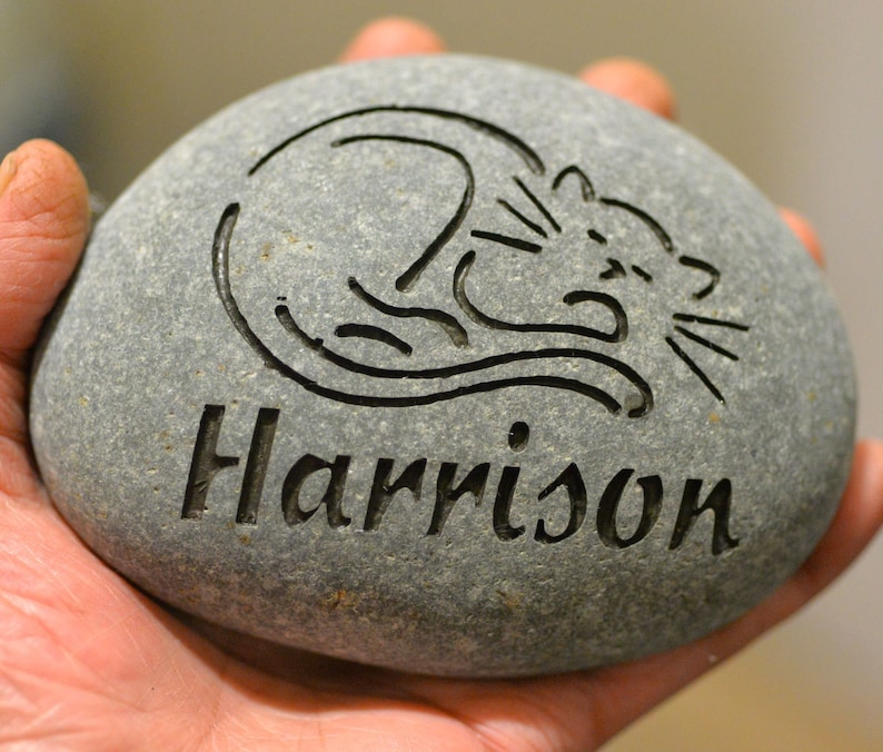 May include: A gray stone with a black etched design of a cat and the name "Harrison" engraved on it.