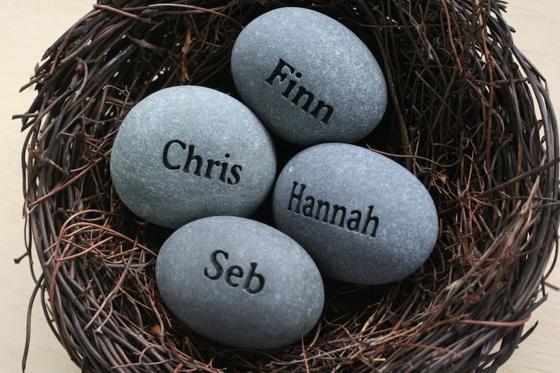 May include: Four grey stones with the names "Finn", "Chris", "Hannah", and "Seb" engraved on them, nestled in a brown woven nest.