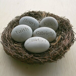 May include: Five grey stones with names engraved on them, arranged in a bird's nest. The names are Henry, Faith, Kaleb, Gabrielle, and Bridget.