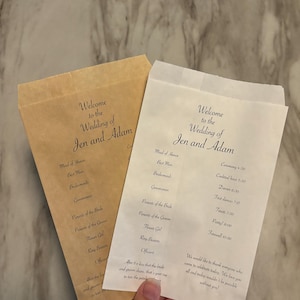 Pode incluir: Duas sacolas de papel brancas com o texto "Bem-vindos ao casamento de Jen e Adam" impresso nelas. A sacola à direita tem um cronograma de casamento impresso, incluindo os horários da cerimônia, do coquetel, do jantar e da despedida.
