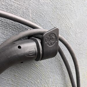 Può includere: Primo piano di un cavo elettrico nero con un gancio in plastica nera. Il gancio ha un logo in rilievo con il testo "Marine" e un simbolo.