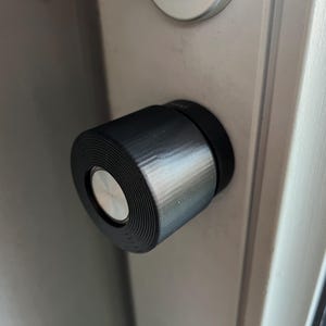 May include: A close-up of a black door handle with a silver ring around the centre. The handle is mounted on a grey door.