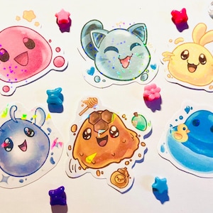 Puede incluir: Una colección de pegatinas coloridas y de estilo caricaturesco que representan personajes sonrientes con forma de mancha en rosa, azul, amarillo y marrón. Adornos decorativos en forma de estrella y gema rodean las pegatinas.