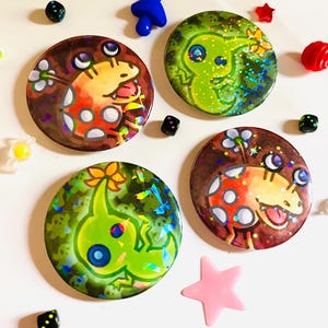 Könnte beinhalten: Vier runde, farbenfrohe Ansteckbuttons mit cartoonartigen Illustrationen von Kreaturen. Die Buttons weisen Farbtöne von Braun, Orange, Grün und Weiß auf. Kleine, farbenfrohe dekorative Formen, darunter Sterne, Würfel und ein Pilz, sind verstreut.