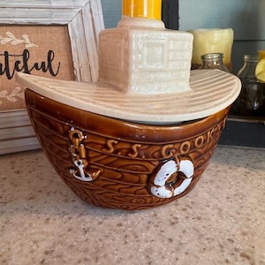 May include: A brown ceramic boat-shaped cookie jar with a cream-colored lid. The jar features an anchor and life preserver design. The lid has a yellow chimney. The text "S.S. COOKIE" is visible on the side.