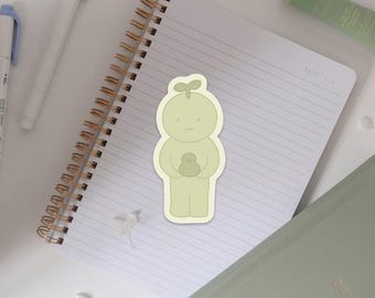 May include: A light green sticker featuring a cartoon character with a sprout on its head, holding a smaller object. The sticker is on a spiral-bound notebook with lined paper, alongside a pen and a tube of lotion.