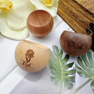 May include: Three wooden bowls in varying shades of brown. One bowl has a floral design and the name "Ruth" engraved. Another bowl has the name "Ruth" engraved. The bowls are on a white surface with green leaves and a wooden box.