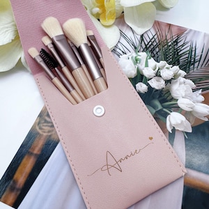 Può includere: Un set di pennelli per il trucco in un astuccio rosa. I pennelli hanno setole chiare e manici metallici. L'astuccio è personalizzato con il nome "Annie" in corsivo e un piccolo simbolo a forma di cuore.