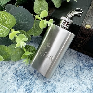 Puede incluir: Una petaca de acero inoxidable plateada con un diseño floral y la palabra "Faith" grabada en la parte delantera. La petaca tiene una tapa plateada y una cadena plateada con un cierre plateado.