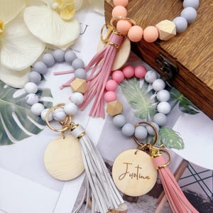 May include: Keyrings with silicone beads in shades of grey, white, pink, and peach. Each keyring features a wooden tag, a tassel, and a gold-coloured clasp. One keyring has the name "Justina" engraved on the wooden tag.