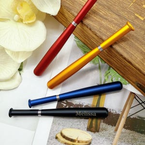 Könnte beinhalten: Vier Baseballschläger-förmige Stifte in Rot, Gold, Blau und Schwarz. Der schwarze Stift hat den eingravierten Namen "Melissa". Jeder Stift hat ein silbernes Band in der Mitte. Die Stifte sind auf einer weißen Oberfläche mit einem Holztablett und floralen Akzenten angeordnet.