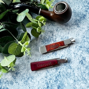 Puede incluir: Dos navajas de bolsillo personalizadas con mangos de madera. La navaja de arriba tiene el nombre "Mathew" grabado. La navaja de abajo tiene el nombre "Garrett" grabado.