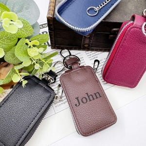 Pode incluir: Porta-chaves de couro em várias cores, incluindo preto, castanho, bordô e azul. O porta-chaves castanho é personalizado com o nome "John". Cada porta-chaves tem um fecho de correr e um porta-chaves de metal.