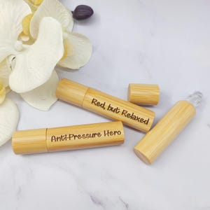 Könnte beinhalten: Drei Bambus-Rollerflaschen für ätherische Öle mit eingraviertem Text. Eine Flasche trägt die Aufschrift "Red, but Relaxed", eine andere "Anti-Pressure Hero". Eine weiße Blume und eine lila Knospe im Hintergrund.