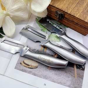 Pode incluir: Descascadores de vegetais em aço inoxidável com acabamento prateado. Um descascador tem o nome "Brooke" gravado na alça. Os descascadores estão dispostos em uma superfície decorativa perto de uma caixa de madeira e flores brancas.