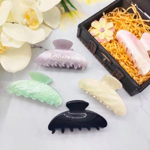 以下が含まれることがあります： ラベンダー、グリーン、クリーム、ブラックなど、さまざまな色のヘアクリップのコレクション。クリップの1つには「Kristina」という名前が刻まれています。クリップは白い表面に配置され、背景には装飾的な箱があります。
