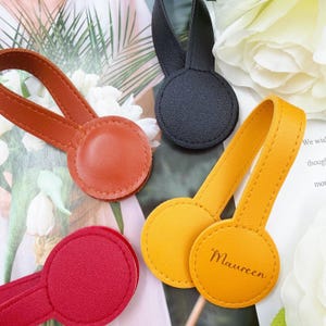 May include: A collection of colorful, round leather curtain tiebacks in shades of red, orange, yellow, and black. Each tieback features a loop and a circular base, with one displaying the name "Maureen".