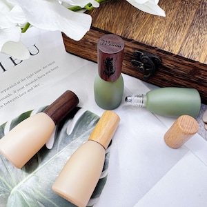 Puede incluir: Varias botellas cosméticas pequeñas y vacías con tapas de madera dispuestas sobre una superficie blanca. Las botellas son de tonos beige y verde. Una botella tiene una tapa de madera con el nombre "Monica" grabado.