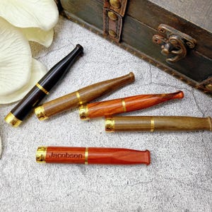 May include: A collection of wooden cigarette holders in various shades of brown and red, accented with gold-colored metal bands. One holder is engraved with the name "Jacobson". The holders are arranged near a wooden chest.