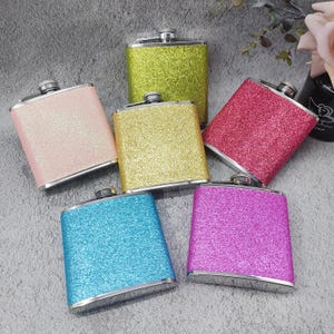 May include: Six different coloured, glitter-covered, metal hip flasks. The flasks are arranged in a semi-circle on a grey surface. The colours are pink, gold, red, purple, blue, and green.