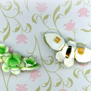 May include: Two floral hair accessories. One is a cluster of green and white flowers with green leaves. The other is a white flower with a black and gold center and amber accents. The background is a floral pattern.