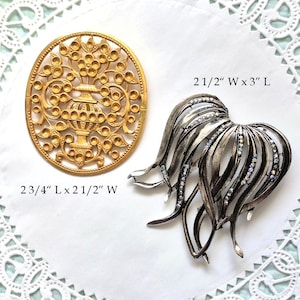 May include: An oval gold-tone brooch featuring a central urn and floral motif measuring 2 3/4 inches long by 2 1/2 inches wide, and a silver-tone brooch with curved, flowing ribbons accented by small iridescent crystals measuring 2 1/2 inches wide by 3 inches long.