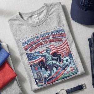 以下が含まれることがあります： 「WORLD CUP 2026 WELCOME TO AMERICA」という文字がプリントされたヘザーグレーのTシャツ。デザインには、サッカーボールを蹴る自由の女神像、アメリカ国旗、ゴールデンゲートブリッジ、都市のスカイラインが描かれています。