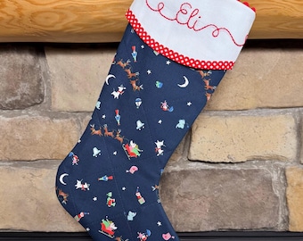 May include: A navy blue quilted Christmas stocking featuring a repeating pattern of reindeer, sleighs, and stars. The white cuff is embroidered with the name Eli in red cursive script and is trimmed with red and white polka dot fabric and red pom-poms.
