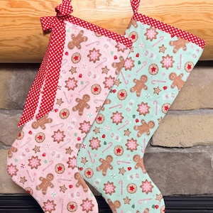 May include: Two Christmas stockings, one light pink and one light green, featuring a repeating pattern of gingerbread men, star-shaped cookies, and candy canes. Both stockings have a red and white polka dot fabric trim and a matching hanging loop.