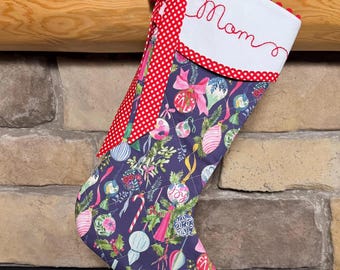 Peut inclure: Une chaussette de Noël matelassée en tissu bleu foncé avec un motif coloré d'ornements, de rubans et de feuillage. Le revers supérieur est blanc avec le mot Mom brodé en rouge, agrémenté d'un nœud et d'une bordure à carreaux vichy rouges.