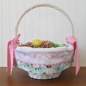 May include: White wicker Easter basket with a pink and white polka dot ribbon, a white fabric liner with pink stitching, and a floral print fabric trim. The basket is filled with pastel colored Easter eggs and shredded paper.