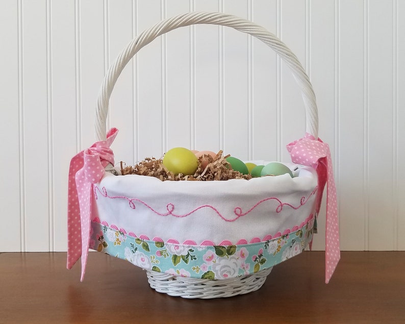 May include: White wicker Easter basket with a pink and white polka dot ribbon, a white fabric liner with pink stitching, and a floral print fabric trim. The basket is filled with pastel colored Easter eggs and shredded paper.