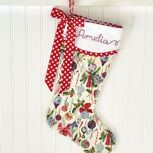 May include: A white Christmas stocking with a red and white gingham ribbon, a white cuff with the name "Amelia" embroidered in red, and a colorful print of ornaments and candy canes.