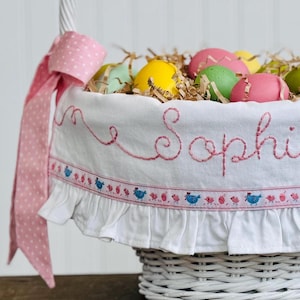 May include: A white wicker Easter basket with a pink polka dot bow and handle. The basket contains colorful eggs and is lined with a white fabric embroidered with the name "Sophie" in pink. A decorative border with blue birds and pink flowers adorns the fabric.