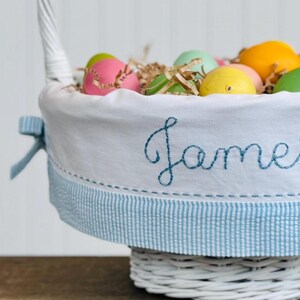 May include: White wicker basket with a blue and white gingham trim and a white fabric liner. The liner has the name "James" embroidered on it in blue thread. The basket is filled with colorful Easter eggs and shredded paper.