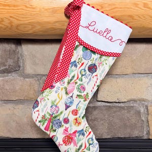 May include: A Christmas stocking featuring a white cuff embroidered with the name Luella in red script. The cuff is trimmed with red and white pom-poms and a red and white gingham loop. The body displays a colorful pattern of ornaments.