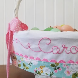 May include: White wicker basket with a pink and white polka dot ribbon, a white fabric liner with pink scalloped trim, and the name "Caro" embroidered in pink thread. The basket is filled with pastel colored Easter eggs.