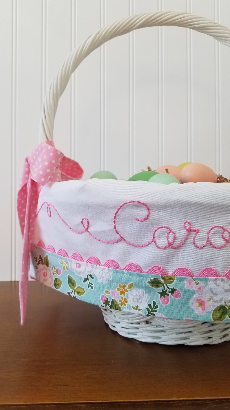 May include: White wicker basket with a pink and white polka dot ribbon, a white fabric liner with pink scalloped trim, and the name "Caro" embroidered in pink thread. The basket is filled with pastel colored Easter eggs.